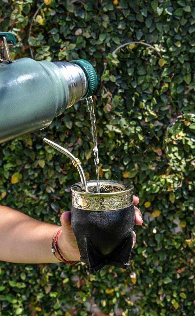 A hand pours hot water into a traditional mate cup against a leafy background.